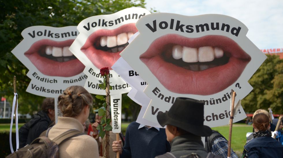 Demostranten mit Protestschildern, auf denen jeweils ein Mund zu sehen ist. Der Schriftzug auf den Plakten lautet: Volksmund. Endlich mündig? Volksentscheid!