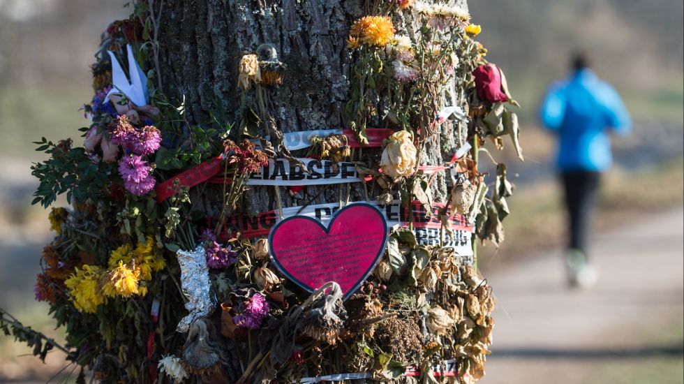 Ein Polizeiabsperrband ist um einen Baum gewickelt. Passanten klemmten in das Absperrband Blumen.