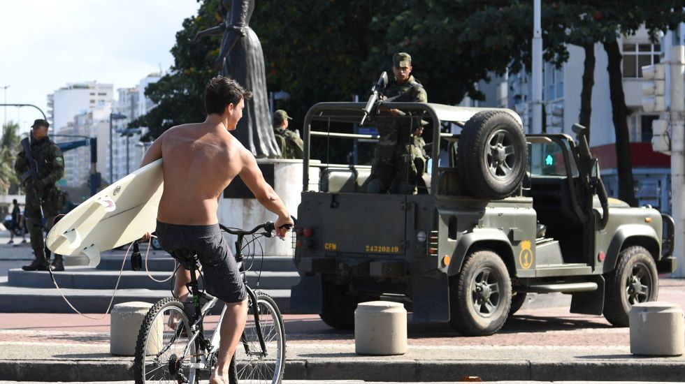 Ein Surfer fährt an der Copacabana von Rio de Janeiro an einem Jeep der Armee mit einem Soldaten, der ein Gewehr in der Hand hält, vorbei