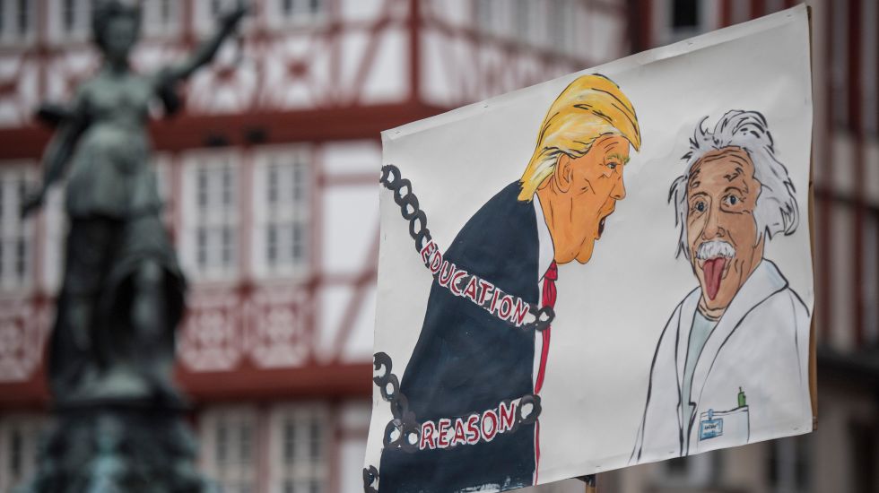 Ein Plakat wird hochgehalten. Darauf abgebildet Albert Einstein, die Zunge hinausstreckend. Neben ihm brüllt ihn Trump an, der in Ketten zurückgehalten wird. Auf den Ketten steht geschrieben: "Education" und "Reason"