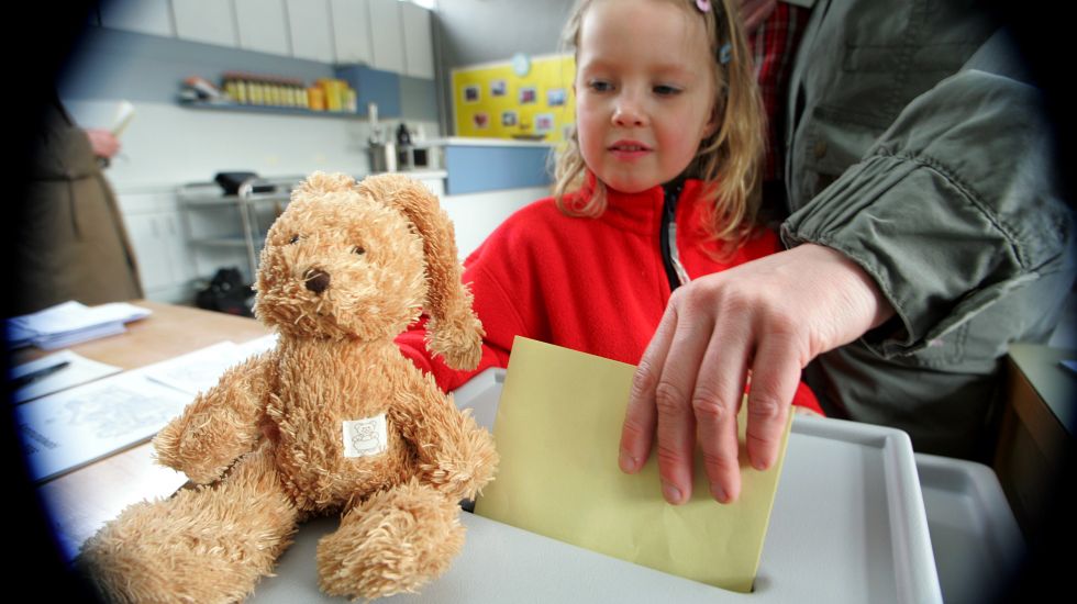 Einwurf eines Stimmzettels in die Wahlurne. Daneben setzt ein Mädchen ihr Stofftier auf die Wahlurne.