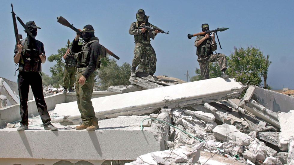 Vier bewaffnete, maskierte Männer stehen auf Trümmern; sonnige Außenaufnahme, Foto.