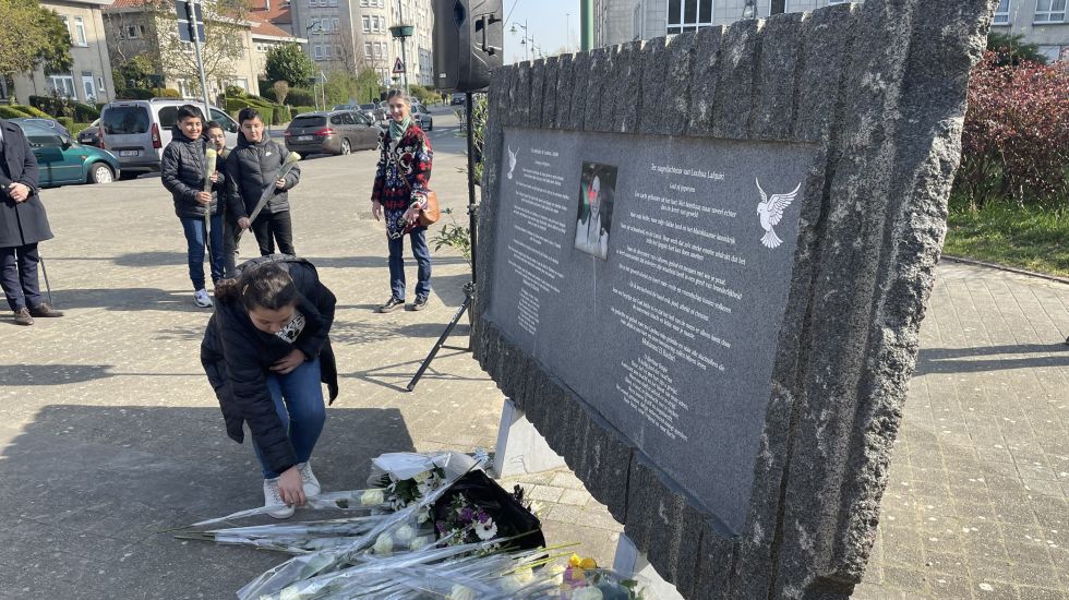 Frau legt Blumen an einer Gedenktafel nieder, mehrere Menschen stehen im Hintergrund, Foto.