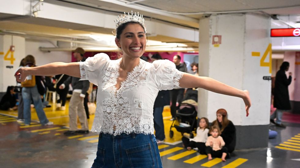 Frau mit Krone und Spitzenbluse tanzt lachend in Tiefgarage; weitere Menschen im Hintergrund. Foto.