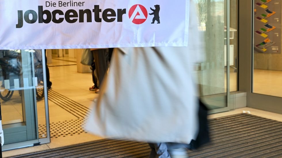 Foto: Person mit hellem Mantel betritt ein Gebäude, darüber Banner mit „jobcenter“-Logo.