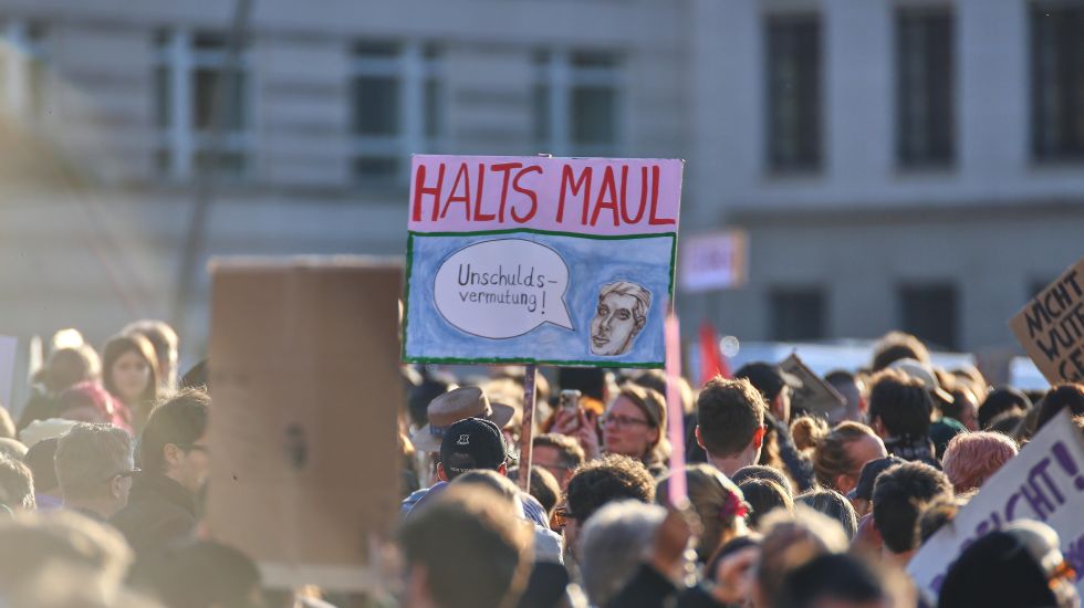 Menschenmenge bei einer Demo, ein Schild mit Zeichnung und Text wird hochgehalten, Foto.