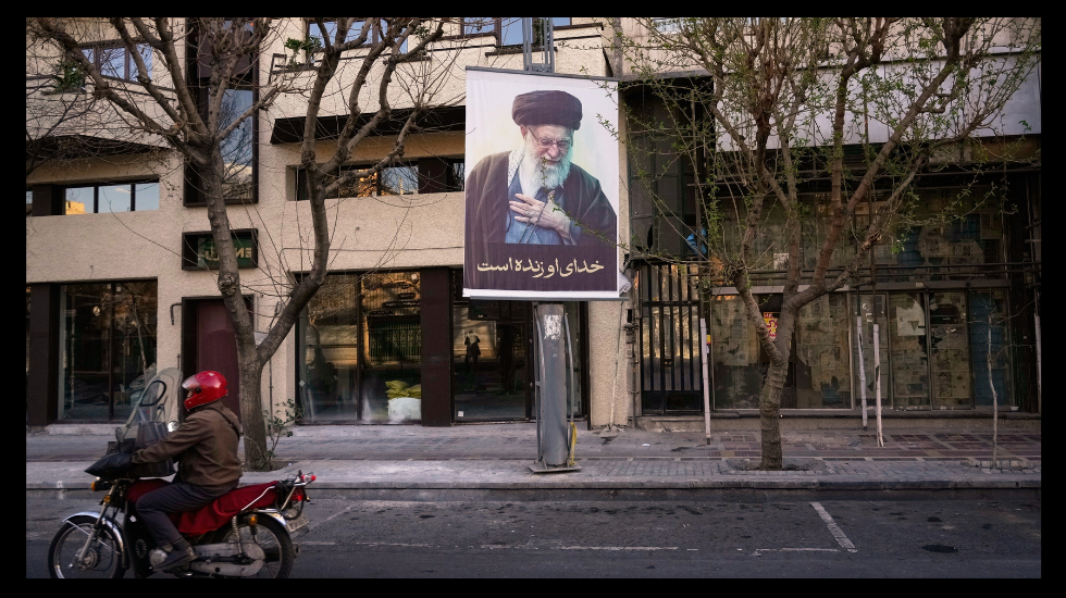 Foto: Motorradfahrer mit rotem Helm fährt an Plakat mit Porträt von Ali Khamenei vorbei.