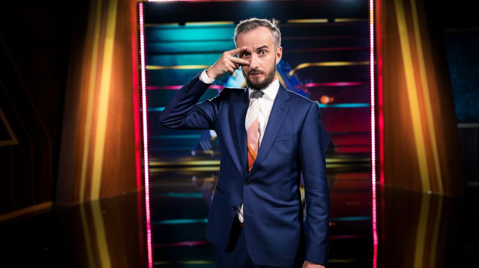 Jan Böhmermann im blauen Anzug vor bunter Lichtwand, posiert mit Hand vor dem Gesicht, Foto.