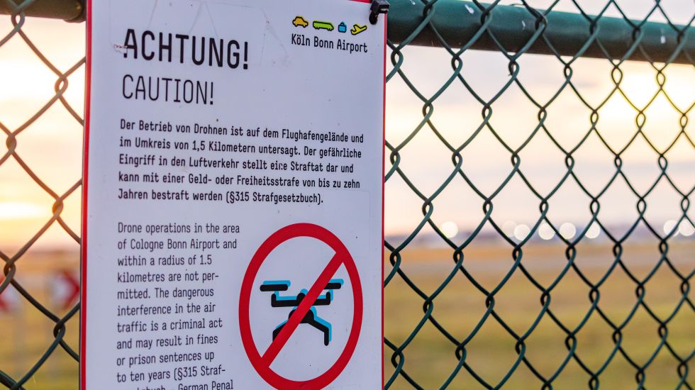Foto eines Warnschilds an einem Zaun, verbietet Drohnen, unscharfer Flughafen im Hintergrund.