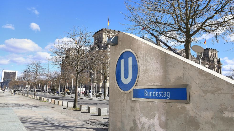 Foto, U-Bahn-Eingang Bundestag mit Reichstagsgebäude im Hintergrund, klare Himmel, Bäume ohne Laub.